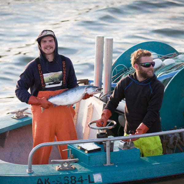 One fishermna holding a freshly-caught salmon next to a fisherman steering the boat | Fresh & Wild | SeaBear Smokehouse